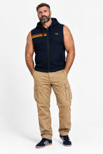 A muscular man wearing a sleeveless navy blue zip-up hoodie with yellow and orange stripes on the chest, tan cargo pants, and navy sneakers, standing against a white studio background.