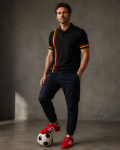 Man in black polo shirt with red and yellow stripes and navy pants standing next to a soccer ball against a gray background.