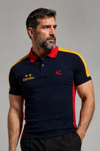 A man wearing a navy blue polo shirt with red and yellow color-blocked details, featuring 'ESPANA' with stars on the right chest and a red 'NC' logo on the left chest, against a plain gray studio background.