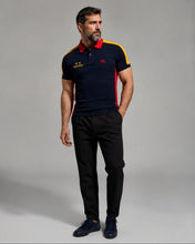 A man modeling a navy blue polo shirt with red and yellow accents, paired with black trousers and navy shoes, against a plain gray studio background.