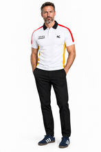 A man modeling a white short-sleeve polo shirt with black, red, and yellow side panels, black collar, and black chest logos, paired with black trousers and navy blue sneakers, against a plain white background.
