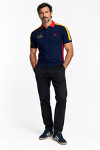 A man modeling a navy blue polo shirt with red and yellow accents, featuring the Spanish flag stars on the right chest and a red 'NC' logo on the left chest, paired with black trousers and navy sneakers.