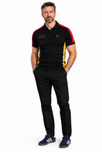 A man modeling a black short-sleeve polo shirt with red and yellow side panels, featuring a red 'GERMANY' text and 'KC' logo on the chest, paired with black trousers and navy blue sneakers, against a plain white background.