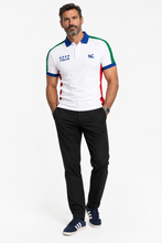 A man modeling a white short-sleeve polo shirt with green, blue, and red trim, featuring 'ITALIA' and 'NC' logos on the chest, paired with black trousers and navy sneakers, against a plain white background.