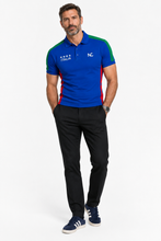A man modeling a short-sleeve polo shirt with a royal blue body and green and red shoulder panels, featuring 'ITALIA' and a star graphic on the chest, paired with black trousers and navy sneakers against a white studio background.