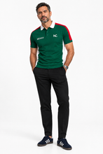 A man modeling a green short-sleeve polo shirt with white and red color-blocked details on the shoulders and sleeves, featuring 'MEXICO' and 'NC' logos on the chest, paired with black trousers and navy sneakers.