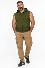 A man modeling a sleeveless olive green zip-up hoodie vest with drawstrings and a small embroidered logo on the chest, paired with tan cargo pants and black sneakers, against a plain white studio background.